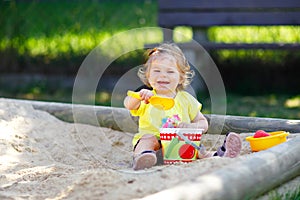 Cute toddler girl playing in sand on outdoor playground. Beautiful baby having fun on sunny warm summer sunny day. Happy