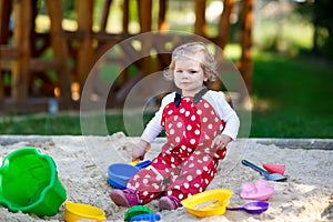 Cute toddler girl playing in sand on outdoor playground. Beautiful baby in red gum trousers having fun on sunny warm