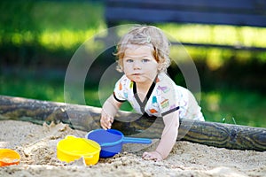 Cute toddler girl playing in sand on outdoor playground. Beautiful baby in red gum trousers having fun on sunny warm