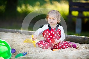 Cute toddler girl playing in sand on outdoor playground. Beautiful baby in red gum trousers having fun on sunny warm