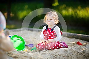 Cute toddler girl playing in sand on outdoor playground. Beautiful baby in red gum trousers having fun on sunny warm