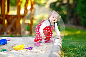 Cute toddler girl playing in sand on outdoor playground. Beautiful baby in red gum trousers having fun on sunny warm