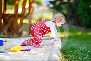 Cute toddler girl playing in sand on outdoor playground. Beautiful baby in red gum trousers having fun on sunny warm