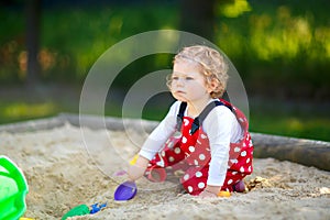 Cute toddler girl playing in sand on outdoor playground. Beautiful baby in red gum trousers having fun on sunny warm