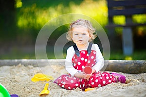 Cute toddler girl playing in sand on outdoor playground. Beautiful baby in red gum trousers having fun on sunny warm