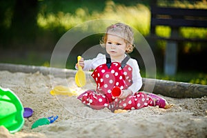 Cute toddler girl playing in sand on outdoor playground. Beautiful baby in red gum trousers having fun on sunny warm