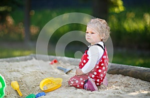 Cute toddler girl playing in sand on outdoor playground. Beautiful baby in red gum trousers having fun on sunny warm