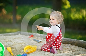 Cute toddler girl playing in sand on outdoor playground. Beautiful baby in red gum trousers having fun on sunny warm