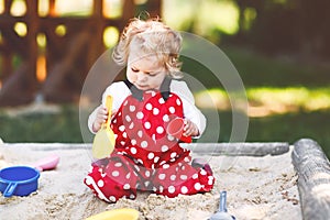 Cute toddler girl playing in sand on outdoor playground. Beautiful baby in red gum trousers having fun on sunny warm