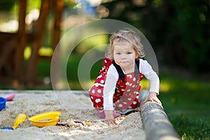 Cute toddler girl playing in sand on outdoor playground. Beautiful baby in red gum trousers having fun on sunny warm