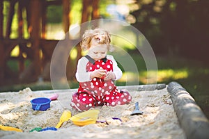 Cute toddler girl playing in sand on outdoor playground. Beautiful baby in red gum trousers having fun on sunny warm