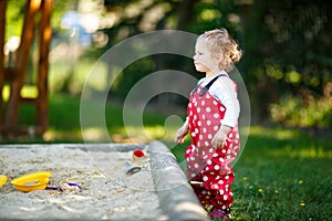 Cute toddler girl playing in sand on outdoor playground. Beautiful baby in red gum trousers having fun on sunny warm