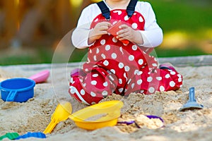 Cute toddler girl playing in sand on outdoor playground. Beautiful baby in red gum trousers having fun on sunny warm