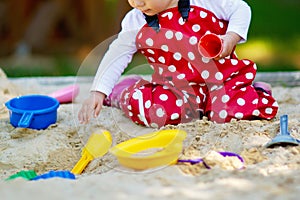 Cute toddler girl playing in sand on outdoor playground. Beautiful baby in red gum trousers having fun on sunny warm