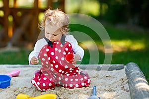 Cute toddler girl playing in sand on outdoor playground. Beautiful baby in red gum trousers having fun on sunny warm