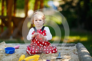 Cute toddler girl playing in sand on outdoor playground. Beautiful baby in red gum trousers having fun on sunny warm