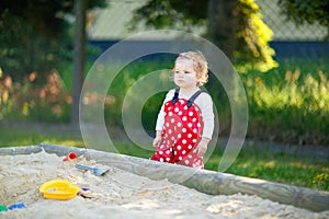 Cute toddler girl playing in sand on outdoor playground. Beautiful baby in red gum trousers having fun on sunny warm