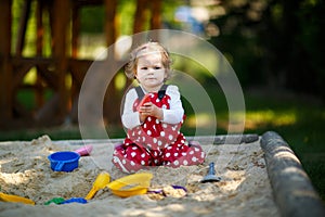 Cute toddler girl playing in sand on outdoor playground. Beautiful baby in red gum trousers having fun on sunny warm