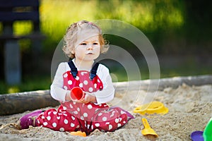 Cute toddler girl playing in sand on outdoor playground. Beautiful baby in red gum trousers having fun on sunny warm