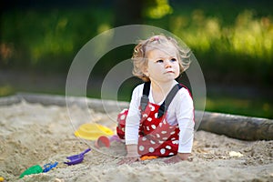 Cute toddler girl playing in sand on outdoor playground. Beautiful baby in red gum trousers having fun on sunny warm