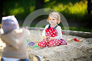 Cute toddler girl playing in sand on outdoor playground. Beautiful baby in red gum trousers having fun on sunny warm
