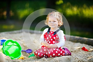 Cute toddler girl playing in sand on outdoor playground. Beautiful baby in red gum trousers having fun on sunny warm