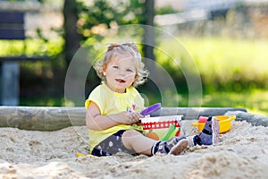 Cute toddler girl playing in sand on outdoor playground. Beautiful baby in red gum trousers having fun on sunny warm