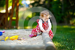 Cute toddler girl playing in sand on outdoor playground. Beautiful baby in red gum trousers having fun on sunny warm