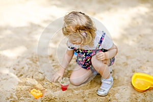 Cute toddler girl playing in sand on outdoor playground. Beautiful baby in red trousers having fun on sunny warm summer