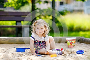 Cute toddler girl playing in sand on outdoor playground. Beautiful baby in red trousers having fun on sunny warm summer