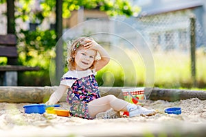 Cute toddler girl playing in sand on outdoor playground. Beautiful baby in red trousers having fun on sunny warm summer
