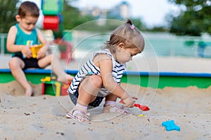 Cute toddler girl playing in sand on outdoor playground. Beautiful baby in red trousers having fun on sunny warm summer day. Child