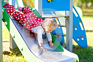 Cute toddler girl playing on slide on outdoor playground. Beautiful baby in red gum trousers having fun on sunny warm