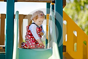 Cute toddler girl playing on slide on outdoor playground. Beautiful baby in red gum trousers having fun on sunny warm
