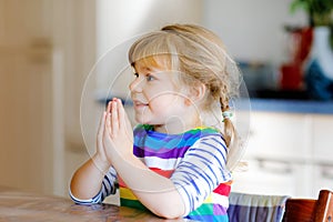 Cute toddler girl praying to God at home. Child using hands for pray and thank for food. Christian tradition.
