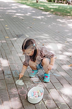 Cute toddler girl sitting and drawing with chalk on asphalt in park