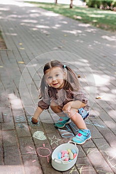 Cute toddler girl sitting and drawing with chalk on asphalt in park. Child holding chalk