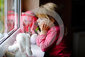 Cute toddler girl sitting by window and looking out on rainy day. Dreaming child with doll and soft toy feeling happy