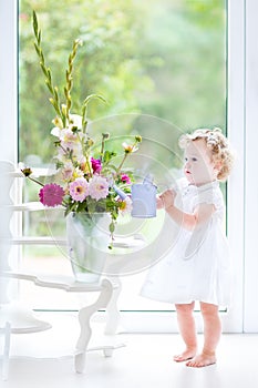 Cute toddler girl in white dress watering flowers