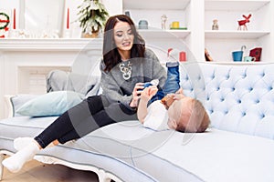 Cute toddler lies on the blue couch and happy young mommy kneading his legs and makes him happy.