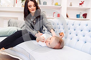 Cute toddler lies on the blue couch and happy young mommy kneading his legs and makes him happy.