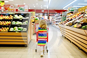 Cute todler girl pushing shopping cart in supermarket. Little child buying fruits. Kid grocery shopping. Adorable baby