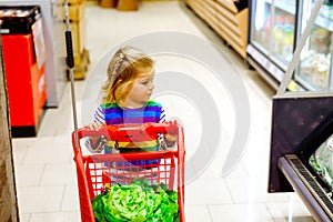 Cute todler girl pushing shopping cart in supermarket. Little child buying fruits. Kid grocery shopping. Adorable baby