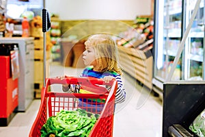 Cute todler girl pushing shopping cart in supermarket. Little child buying fruits. Kid grocery shopping. Adorable baby