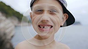 A cute toothless boy in cap is smiling on the camera at sea background. Portrait of a smile schoolboy without teeth