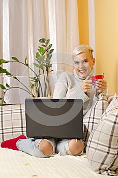 Cute trendy girl sitting on the couch with coffee and laptop