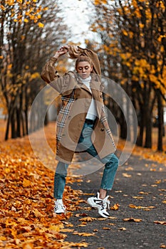 Cute trendy young woman blonde woman straightens hair. Attractive girl in a stylish coat in a sweater in jeans in sneakers posing