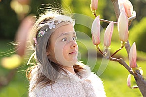 Cute tween girl with with handmade wreath on head with funny face expression looking surprisingly at magnolia tree buds.