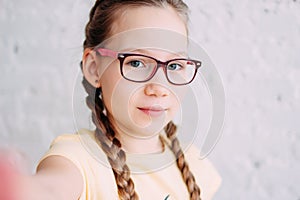 Cute tweens girl in yellow t-shirt with funny pigtails taking selfie on frontal camera on the white brick wall background