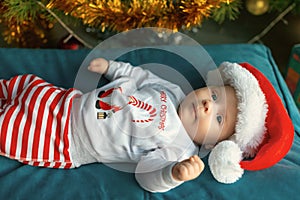 Cute two-month baby in a striped pants and santa hat lying under Christmas tree and looking at camera. Caucasian infant